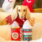 Dog wearing a red sweater with Christmas-themed hot chocolate cups and a gingerbread man toy.